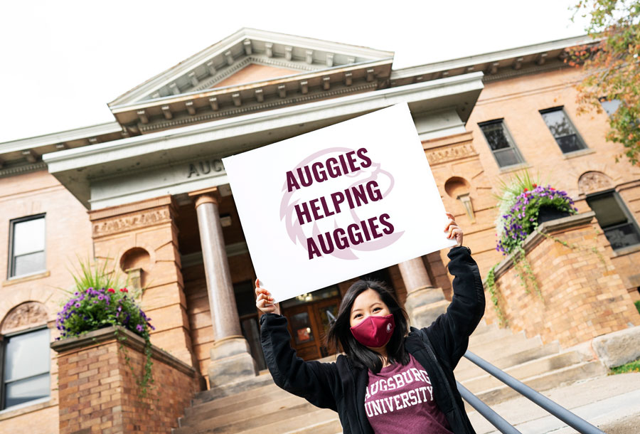 Student wearing a mask is holding a sign that says "Auggies Helping Auggies" on the front steps of Old Main.